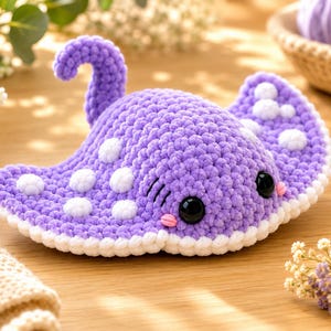 May include: A purple crochet stingray plush toy featuring a textured yarn surface, white polka dot patterns on its wings, a curved tail, large black plastic safety eyes, and small pink stitched cheeks, finished with a white border along the edges.