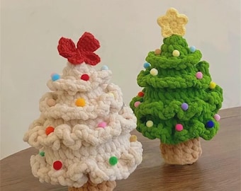 May include: Two crocheted Christmas tree ornaments made from thick yarn. One tree is cream-coloured with a red bow on top, and the other is green with a yellow star on top. Both trees are decorated with small, colourful pom-poms and have brown trunks.