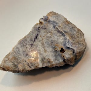Könnte beinhalten: Eine dreieckige Platte aus poliertem Mineral mit einer durchscheinenden weißen und grauen Basis, durch die sich tiefviolette Adern ziehen. Die Oberfläche weist eine glatte, glänzende Textur mit natürlichen Rissen und einer kleinen Vertiefung auf.