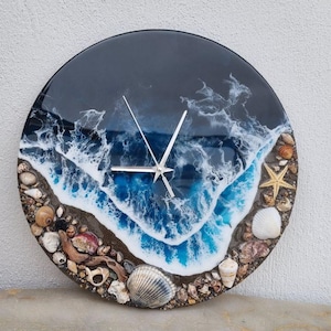 Könnte beinhalten: Runde Wanduhr mit Küstenmotiv. Das Zifferblatt zeigt einen dunkelblauen Ozean mit weißen, schäumenden Wellen und einem sandigen Ufer. Die Uhr ist mit Muscheln, Kieselsteinen und einem Seestern verziert.