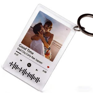 Puede incluir: Llavero acrílico transparente con una foto de una pareja en la playa. El diseño incluye una interfaz de reproductor de música con el título de la canción "Good Time" de Owl City, Carly Rae Jepsen. Un llavero plateado está adjunto.