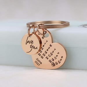 May include: A rose gold keychain with a heart-shaped calendar pendant and a round pendant. The round pendant says "love you". The heart-shaped pendant displays a calendar for June 2027.