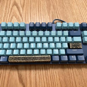 May include: A mechanical keyboard featuring a color scheme of light blue and dark blue keys. The space bar and the right shift key are replaced with metallic, gold-toned custom keycaps that feature intricate, embossed, ancient-style patterns.