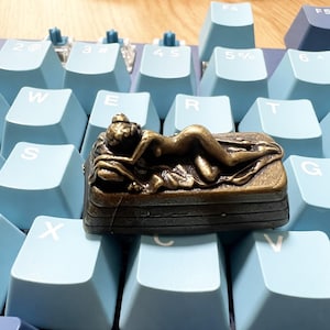 Può includere: Un tasto artigianale color bronzo con un rilievo scolpito di una figura distesa su una base rettangolare. La dettagliata statuetta metallica è posizionata sopra una tastiera meccanica azzurra con lettere bianche sui tasti.