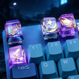 May include: Four custom artisan keycaps featuring a translucent purple resin finish with embedded gold-colored celestial motifs including stars, a crescent moon, a planet with rings, and a rocket ship, installed on a light blue mechanical keyboard.