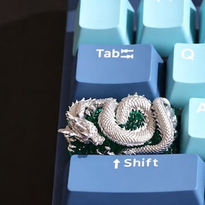 May include: A custom mechanical keyboard keycap featuring a detailed silver-coloured dragon coiled over a translucent emerald green base. The keycap is positioned between a blue Tab key and a blue Shift key on a light blue and blue keyboard.