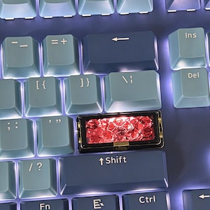 Puede incluir: Un teclado mecánico con teclas de color azul claro y azul oscuro con letras blancas. Una tecla rectangular personalizada con un marco negro y un inserto rojo translúcido facetado se encuentra sobre la tecla de mayúsculas derecha.