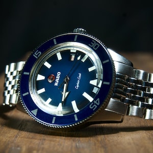 May include: A stainless steel Rado Captain Cook watch with a blue dial and bezel. The watch features white hour markers and hands, and a stainless steel bracelet. The watch is displayed on a wooden surface.