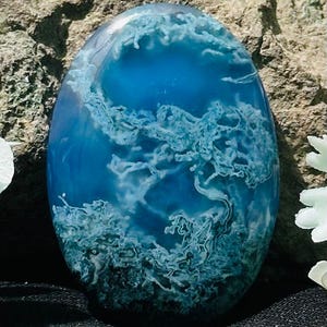 May include: Oval-shaped blue and white stone with a swirling pattern resembling ocean waves. The stone has a polished, glossy finish and is set against a natural rock background. The stone's color transitions from a deep blue to a lighter shade.