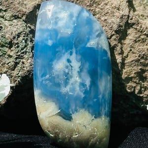 Può includere: Una gemma oblunga lucidata con un sorprendente motivo blu e bianco che ricorda le nuvole nel cielo. La parte inferiore passa a un colore beige e marrone chiaro, creando un contrasto naturale e terroso. La pietra è liscia e riflettente.
