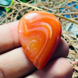 May include: A polished, teardrop-shaped carnelian gemstone featuring a smooth, glossy surface with vibrant orange and reddish-orange hues, displaying natural swirling white banding patterns throughout the translucent stone structure.