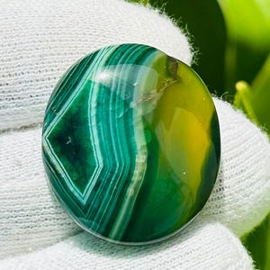 Puede incluir: Una piedra preciosa pulida y redonda con una vibrante mezcla de tonos verdes y amarillos. La piedra presenta un llamativo patrón de bandas concéntricas y un diseño central en forma de diamante. La piedra preciosa se sostiene sobre un paño blanco.