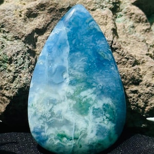 Könnte beinhalten: Ein tropfenförmiger Edelstein mit einer Mischung aus blauen und grünen Farbtönen. Die Oberfläche des Steins weist ein wolkiges Muster auf, mit helleren Blautönen oben und Übergängen zu Grün an der Basis. Der Edelstein ist vor einem strukturierten, braunen Hintergrund platziert.