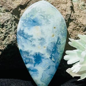 Può includere: Una gemma lucida a forma di goccia con un aspetto nuvoloso e marmorizzato. La pietra presenta una miscela di blu chiaro, bianco e sfumature di verde, che ricorda un acquerello. La gemma è posta su uno sfondo marrone strutturato.