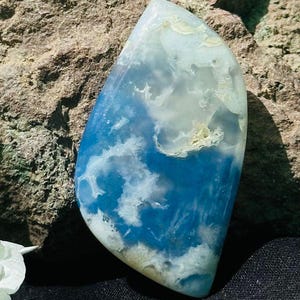 Può includere: Una gemma lucida e allungata con un sorprendente motivo blu e bianco, che ricorda le nuvole nel cielo. La pietra ha una superficie liscia e lucida e una forma naturale e organica. I colori si fondono, creando un design unico e accattivante.
