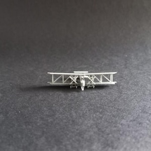 Könnte beinhalten: Ein kleines, hellgraues Doppeldecker-Modell mit zwei Tragflächen und einem detaillierten Rahmen. Das Modellflugzeug hat einen Propeller und ein kleines Cockpit. Die Tragflächen sind durch Streben verbunden, das Design erinnert an die frühe Luftfahrt.