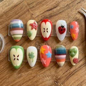 May include: A collection of ten artificial nails with various designs. The designs include colorful stripes, a red star, a red apple, a ladybug, and blue and yellow stars. The nails are displayed on a wooden surface.