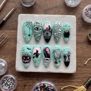 May include: A set of ten almond-shaped press-on nails in a light mint green base color, featuring detailed black and pink Halloween-themed illustrations including a spider web, mummy, gate, skulls, cauldron, pumpkin, witch, coffin, book, and hands.