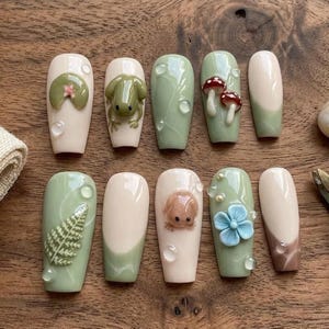 May include: A set of ten artificial nails with a nature-inspired design. The nails feature a variety of designs, including a frog, mushrooms, a fern, a bunny, and a blue flower. The nails are painted in shades of green, white, and brown.