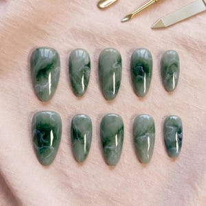 May include: Ten oval-shaped, jade-green press-on nails with a marbled effect. The nails vary in size and are arranged in two rows of five. The nails have a glossy finish and are set against a soft pink background.