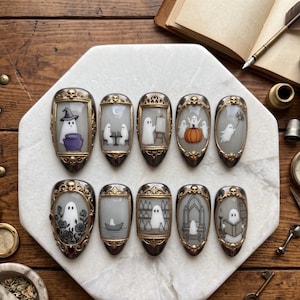 May include: A set of ten press-on nails featuring intricate gold-toned frames with skull accents. Each nail displays a unique hand-painted scene of white ghosts engaged in activities like painting, reading, rowing a boat, and interacting with pumpkins.