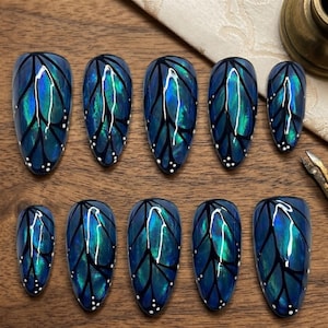 May include: A set of ten artificial nails with a butterfly wing design. The nails are oval-shaped and feature a gradient of blue and teal with black vein-like lines and small white dots. The nails are arranged on a wooden surface.