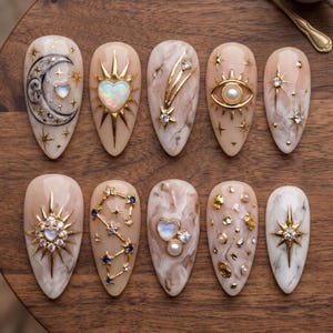 May include: A set of ten almond-shaped press-on nails featuring a beige and white marble base. Designs include gold celestial motifs, sunbursts, crescent moons, heart-shaped opals, pearls, rhinestones, and star constellations on a smooth finish.