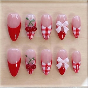May include: A set of ten artificial nails with a variety of designs. Some nails feature a red and white gingham pattern, others have a red French tip, and some are adorned with white bows or cherry accents. The nails are displayed on a light wooden surface.