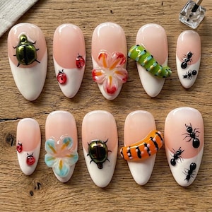 May include: A set of ten artificial nails with various designs. Each nail features a white tip, a pink base, and decorative elements such as ladybugs, flowers, beetles, caterpillars, and ants. The nails are arranged on a wooden surface.