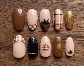 May include: A set of ten oval-shaped press-on nails featuring autumn-themed designs. The collection includes solid brown, black, and gold glitter nails, alongside nude nails decorated with brown plaid patterns, hand-painted pumpkins, and a gold leaf.