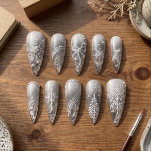 May include: A set of ten almond-shaped press-on nails featuring a shimmering silver-grey base. The designs include white hand-painted snowflakes, pine trees, swirling lines, and small white pearls applied as decorative accents.