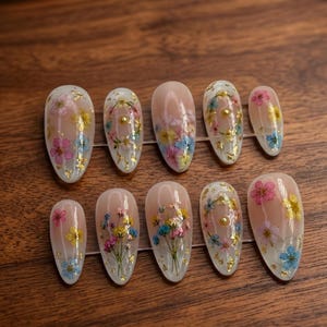 May include: Set of ten artificial nails with a clear base, adorned with dried flowers in pink, yellow, and blue, and gold leaf accents. The nails are oval-shaped and arranged on a wooden surface.