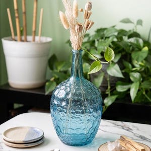 May include: A textured, light blue glass vase filled with dried flowers and seed pods. The vase sits on a white marble surface, alongside small ceramic plates, a spoon, and a wooden coaster with crystals and incense sticks. A potted plant is in the background.