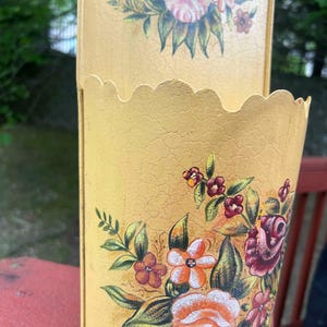 May include: A vintage yellow metal container with a scalloped edge and floral design. The container features a distressed finish and is adorned with hand-painted roses, orange, and burgundy flowers with green leaves. The container is likely used for storage or decoration.