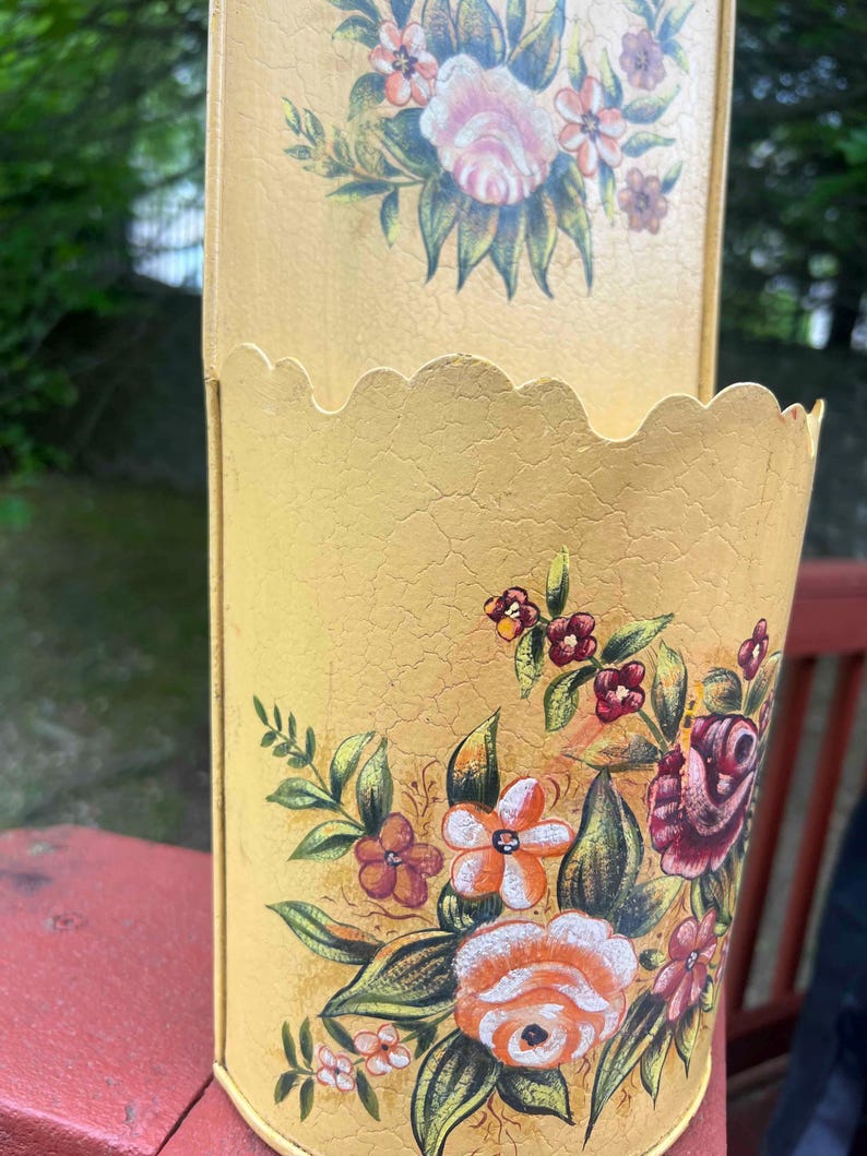 May include: A vintage yellow metal container with a scalloped edge and floral design. The container features a distressed finish and is adorned with hand-painted roses, orange, and burgundy flowers with green leaves. The container is likely used for storage or decoration.