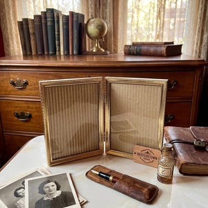 May include: A vintage scene featuring a gold-framed double photo holder, old photographs, a leather pen case with a fountain pen, and a small glass bottle. A wooden dresser and books are in the background.