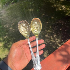 Puede incluir: Un par de cubiertos para ensalada de metal dorado que incluye un tenedor y una cuchara. Cada utensilio tiene un cuenco con un motivo de frutas en relieve y un mango decorado con un patrón de volutas ornamentado y elevado en la base.