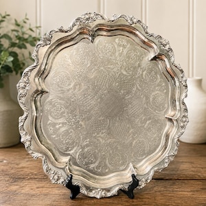 May include: A round silver serving tray featuring an ornate, scalloped edge with detailed floral and scrollwork relief patterns. The center of the tray displays an intricate, etched design consisting of repeating circular motifs and cross-hatching.