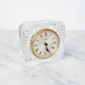 May include: A clear, square quartz clock with a gold-tone bezel and Roman numeral hour markers. The clock face is cream-colored, and the clock is encased in a textured, cut-glass cube. The clock reads "STANGER QUARTZ".