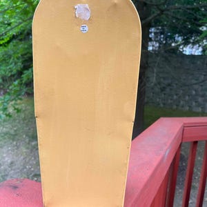 May include: Mustard yellow metal wall decor with a rounded top and a rectangular body. A small sticker on the back reads "Made in India". A small metal hook is attached to the top for hanging.