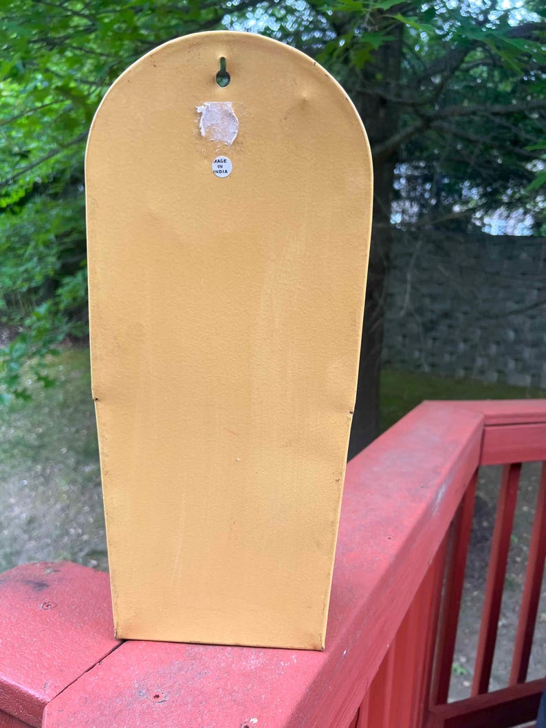 May include: Mustard yellow metal wall decor with a rounded top and a rectangular body. A small sticker on the back reads "Made in India". A small metal hook is attached to the top for hanging.