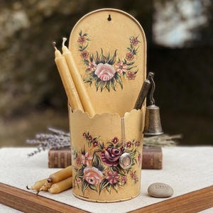 May include: A vintage-style, yellow metal candle holder with a scalloped edge, adorned with hand-painted floral designs in pink, red, and green. The holder contains several long, beige candles and a small bell. A silver locket hangs from the side.