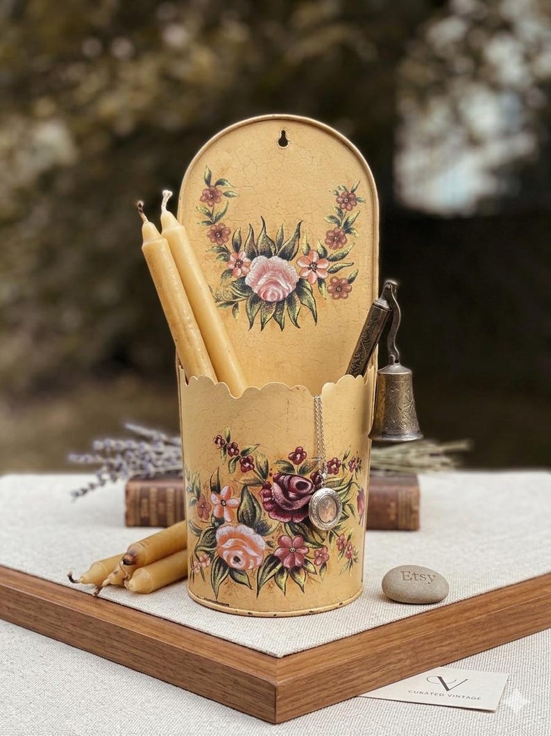 May include: A vintage-style, yellow metal candle holder with a scalloped edge, adorned with hand-painted floral designs in pink, red, and green. The holder contains several long, beige candles and a small bell. A silver locket hangs from the side.