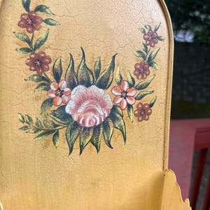 May include: A vintage, wall-mounted metal container with a distressed, yellow finish. The container features a hand-painted floral design in shades of pink, orange, and brown. The container has a scalloped edge and a hook for hanging.