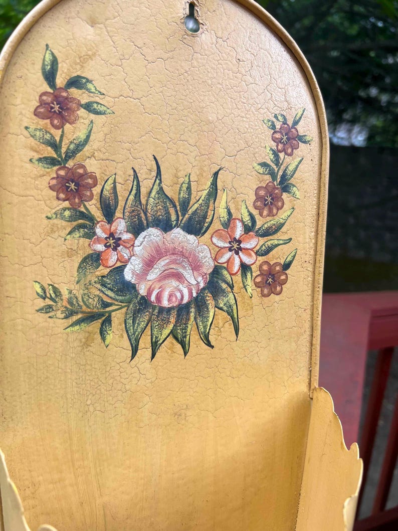 May include: A vintage, wall-mounted metal container with a distressed, yellow finish. The container features a hand-painted floral design in shades of pink, orange, and brown. The container has a scalloped edge and a hook for hanging.