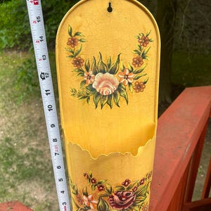 May include: A vintage yellow metal wall pocket with floral designs. The pocket is approximately 16 inches tall and features a scalloped edge. The floral design includes roses and other flowers in shades of pink, red, and brown.