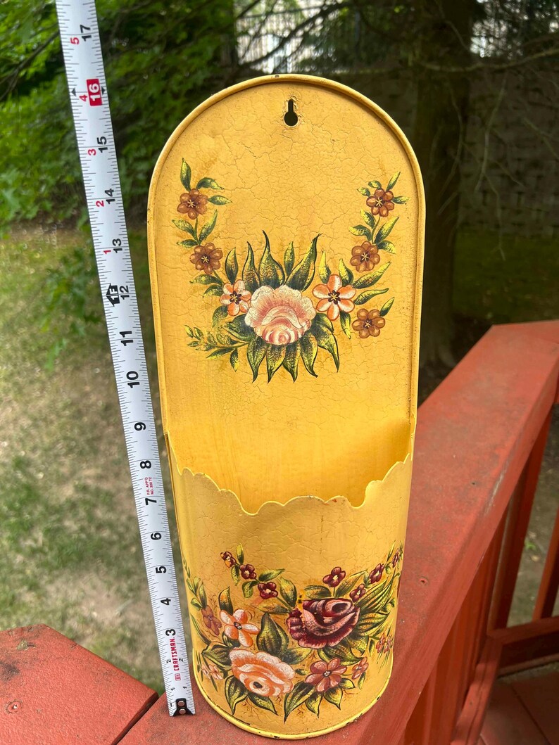 May include: A vintage yellow metal wall pocket with floral designs. The pocket is approximately 16 inches tall and features a scalloped edge. The floral design includes roses and other flowers in shades of pink, red, and brown.