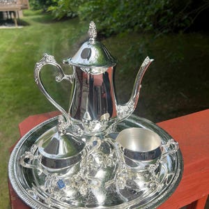 May include: A silver tea set, including a teapot, sugar bowl with lid, creamer, and a serving tray. The set has ornate detailing and is displayed outdoors on a red surface with a green background.
