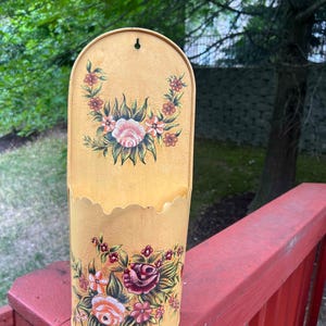 May include: A vintage, yellow metal wall pocket with a scalloped top and floral design. The pocket features hand-painted roses in shades of pink, red, and white, with green leaves. The background has a crackled effect.