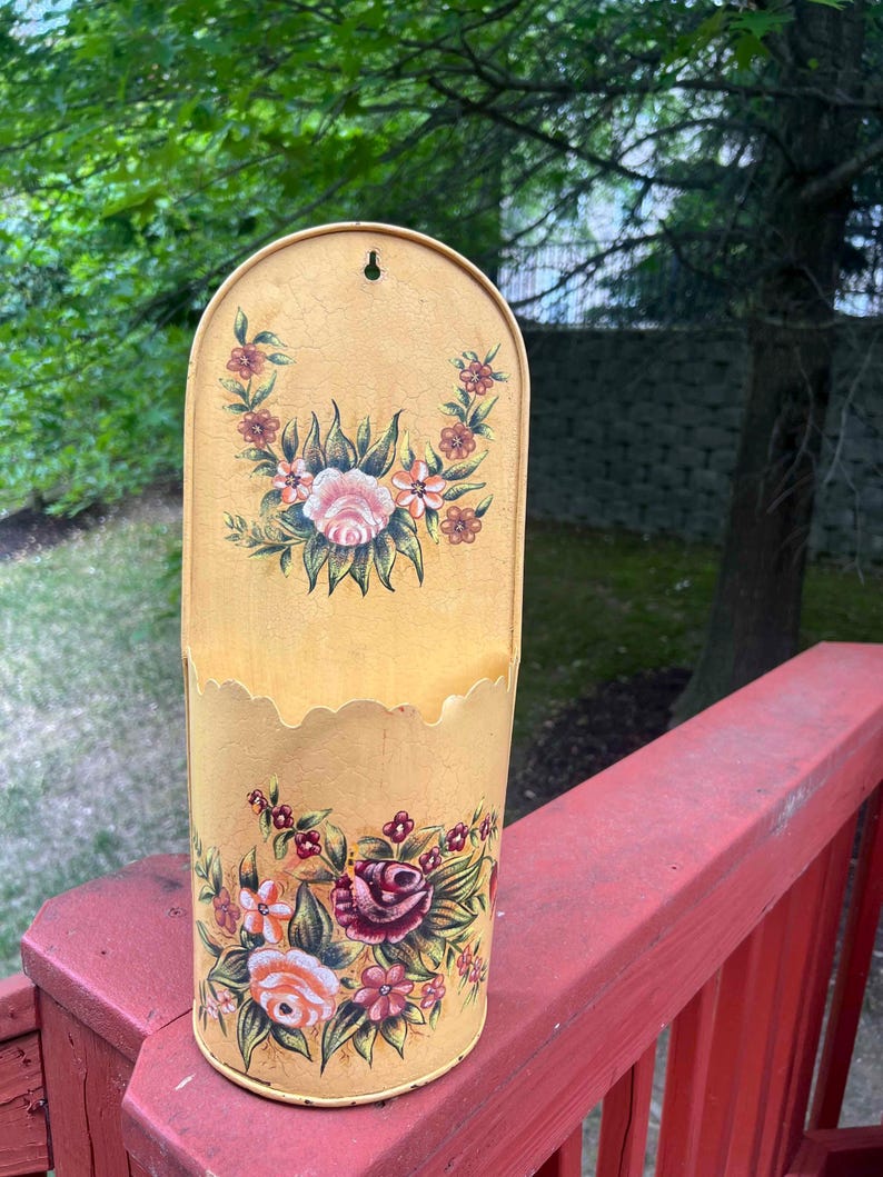 May include: A vintage, yellow metal wall pocket with a scalloped top and floral design. The pocket features hand-painted roses in shades of pink, red, and white, with green leaves. The background has a crackled effect.